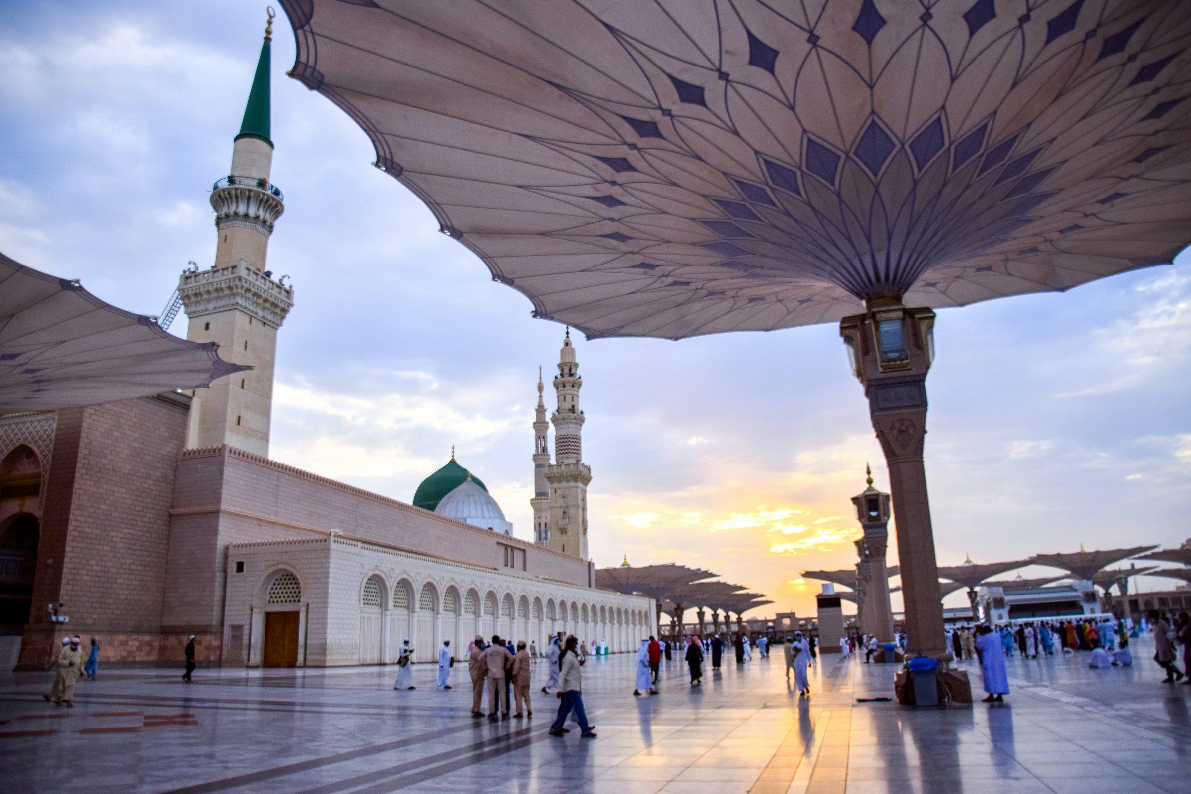 The Green Dome and shade umbrellas of Al-Masjid an-Nabawi at sunset, with people crossing the courtyard